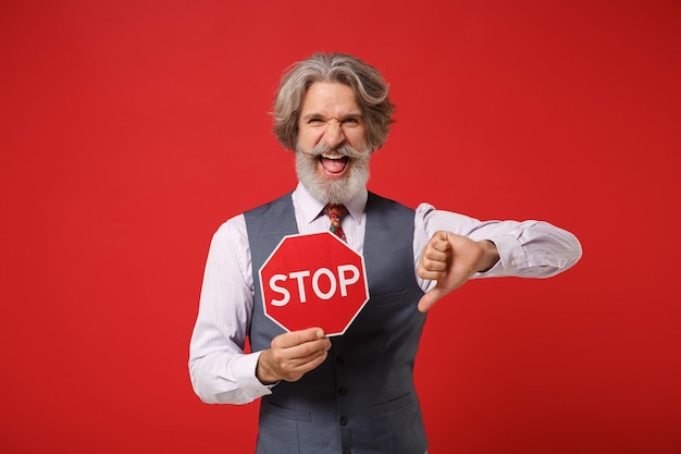 Elderly gray-haired mustache bearded man in classic shirt vest colorful tie isolated on red background. People lifestyle concept. Mock up copy space. Hold red sign with Stop title, showing thumb down.