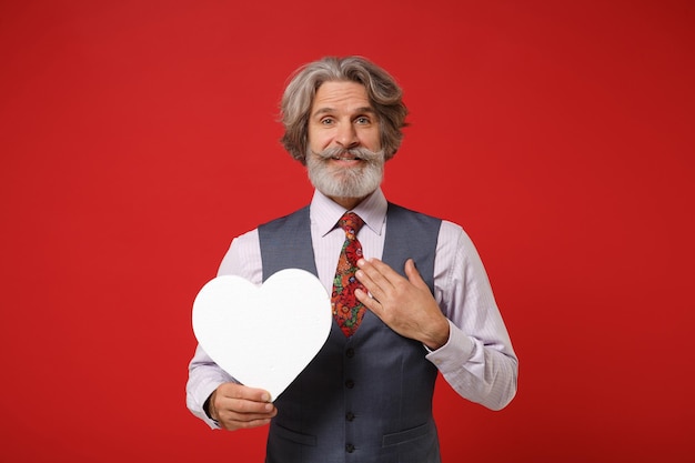 Elderly gray-haired mustache bearded man in classic shirt vest tie posing isolated on red background. People lifestyle concept. Mock up copy space. Holding white blank heart, keeping hand on chest.