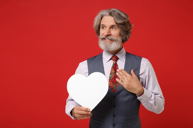 Elderly gray-haired mustache bearded man in classic shirt vest tie posing isolated on red background. People lifestyle concept. Mock up copy space. Holding white blank heart, keeping hand on chest.