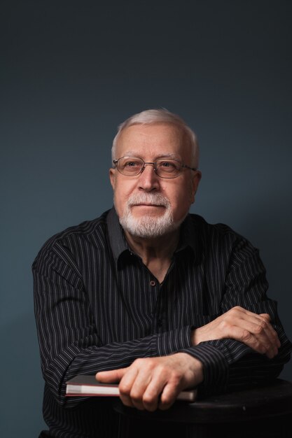 Elderly handsome man sitting down wearing glasses and holding a notebook on dark background