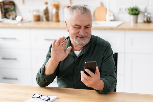 Elderly man communicates online using a mobile phone while sitting at home in the kitchen. Senior man holding smartphone in hands talking on video call