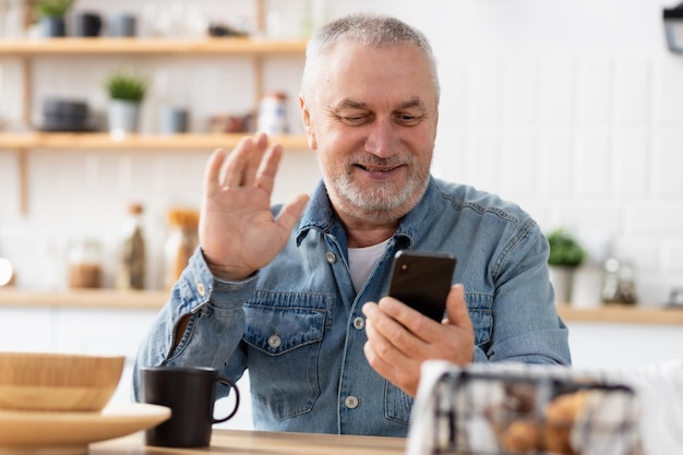 Elderly man communicates online using a mobile phone while sitting at home in the kitchen. Senior man holding smartphone in hands talking on video call