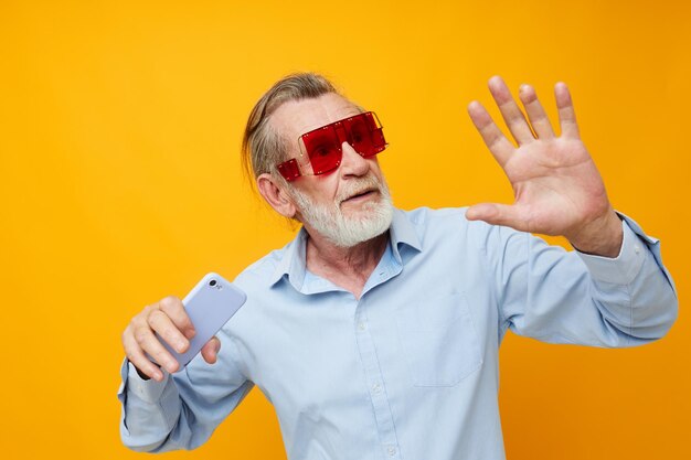 Elderly man in fashionable red glasses with a phone yellow background