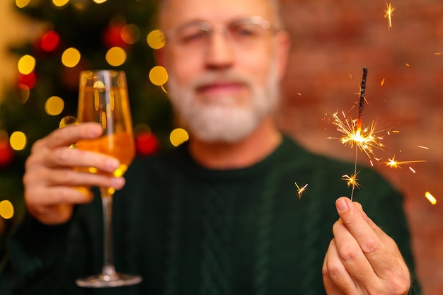 An elderly man in a green knitted sweater raising a toast with champagne near the Christmas tree