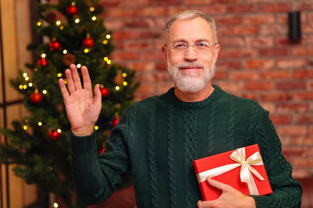 An elderly man in a green knitted with gift box near the Christmas tree