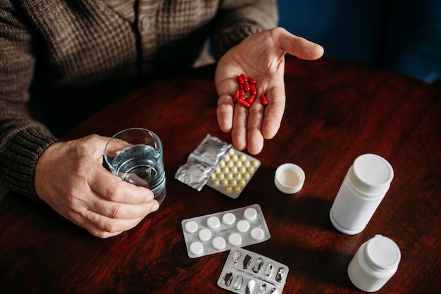 Elderly man hand with pills, home office on background, age-related diseases. Mature senior is ill and being treated in his house