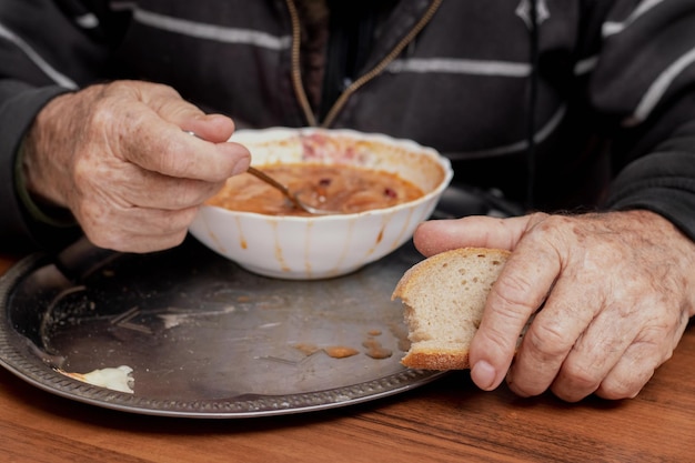 An elderly man is having lunch eating borscht Grandfather holds a spoon and bread near a plate of soup closeup