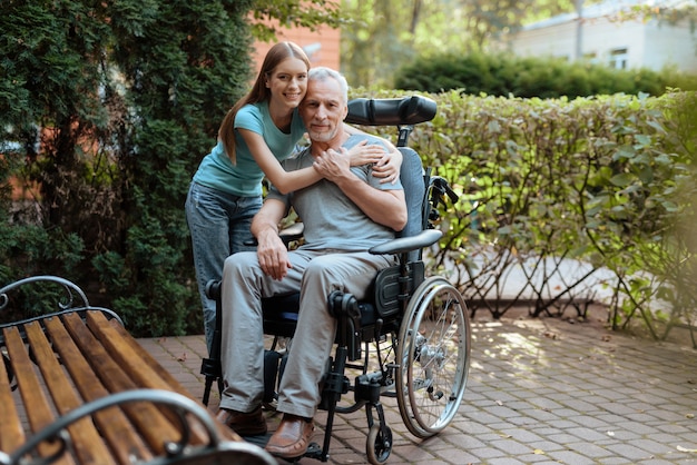 Elderly man is sitting in a wheelchair. Nearby is his daughter and embraces the old man.