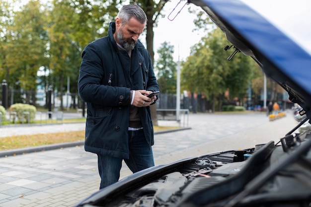 An elderly man next to the raised hood of a broken car calls for technical support on a mobile phone