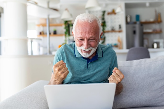 Elderly man seated on couch looking at laptop screen scream with joy feels excited happy celebrating lottery victory lucky moment got online opportunity sales and discounts ecommerce concept