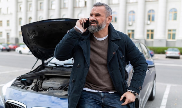 An elderly man sits on the hood of a broken car and calls technical assistance on a mobile phone