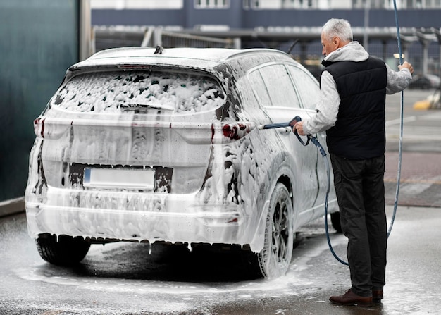 Elderly man spreads cleaning soap suds on his car in manual car wash station Full length portrait