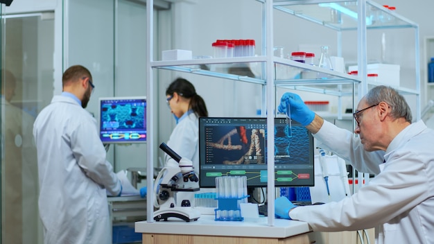 Elderly man technologist doing a laboratory test examining a flask with a blue substance, chemist holding tube with liquids inside. Scientist working with various bacteria tissue and blood samples