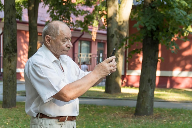 An elderly man walks alone in the park in the summer A modern pensioner businessman in a white shirt and trousers takes pictures with a camera in a mobile phone