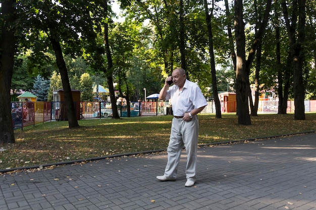 An elderly man walks alone in the park in the summer a modern retired businessman in a white shirt a