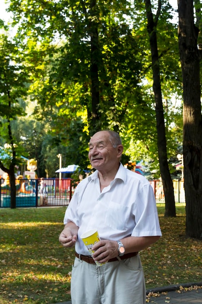 An elderly man walks alone in the park in the summer A pensioner in a white shirt stands alone in the park with a glass of popcorn