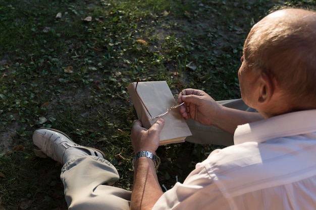 An elderly man in a white shirt is sitting on a blanket on the ground in a park and reading an interesting book A pensioner alone is resting in nature passionate about his hobby