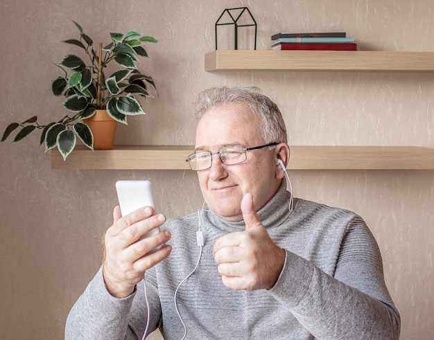 An elderly man with glasses headphones and a smartphone in his hand is studying online courses using a computer Distance learning concept