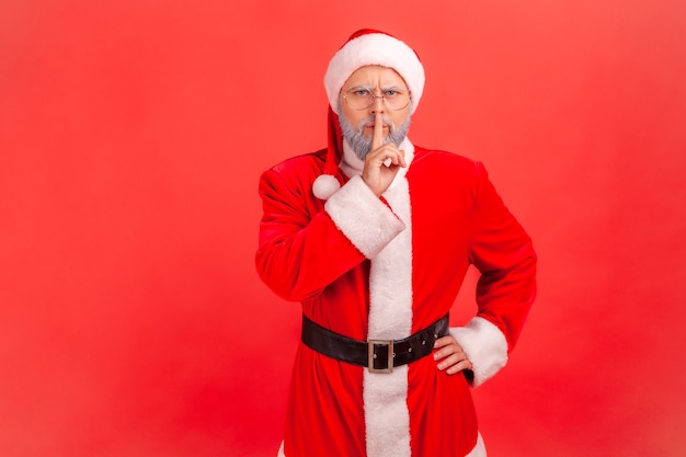 Elderly man with gray beard wearing santa claus costume holding finger on lips making hush silence gesture, asking to keep secret, don't speak. Indoor studio shot isolated on red background.