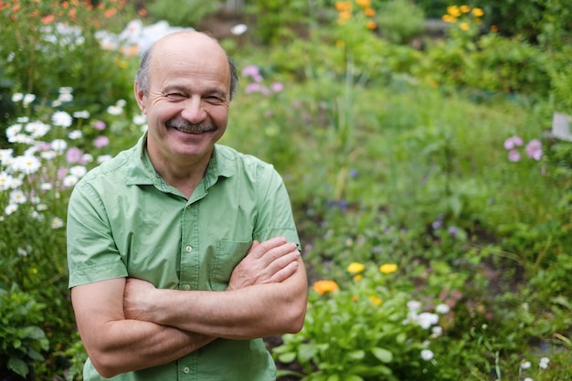 An elderly man with a mustache and a bald spot in a green Tshirt is standing among flowers in the summer garden arms crossed and smiling