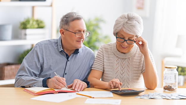 Elderly married couple sitting at the desk with a paper receipt in hands are calculating expenses, managing the family budget.