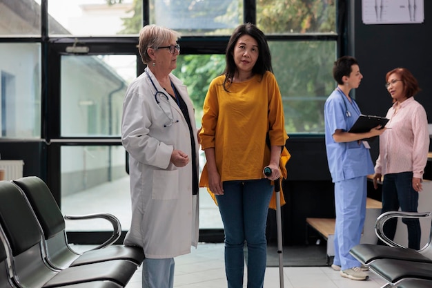 Elderly medic helping injured patient to walk with stick, suffering from physical impairment and using cane. Physician attending physiotherapy to recover from leg injury at facility.