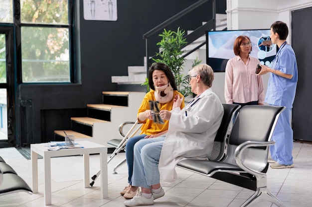 Elderly medic and patient with cervical neck collar looking at xray scan examn, doing checkup visit in hospital waiting room. Asian woman with physical pain and injury having medical appointment.