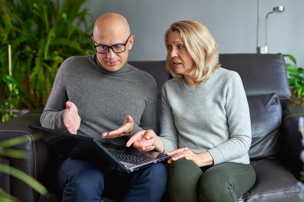 Elderly mother sit rest on sofa with grown-up son watch video on modern laptop