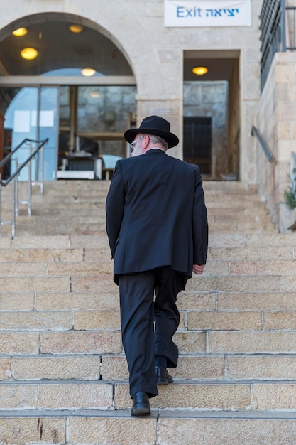 An elderly Orthodox Jew in a black suit and hat walks up the stairs in Jerusalem Vertical Back view