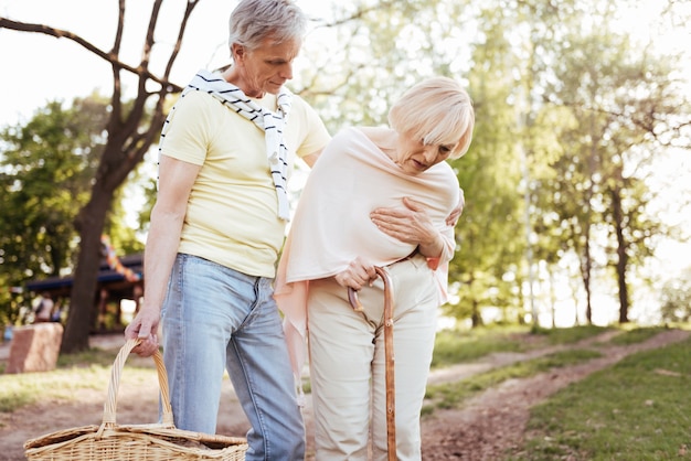 Elderly puzzled sad woman touching her chest and expressing frustration while her husband worrying about her and standing next to her outdoors