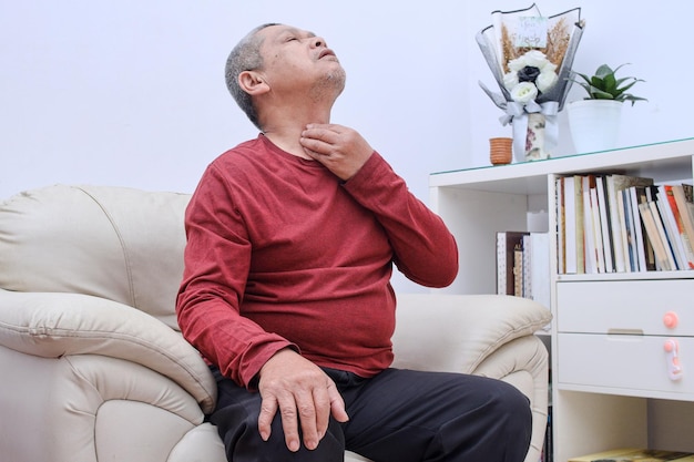 Elderly senior man sitting on sofa touching painful neck sore throat for flu cold and infection