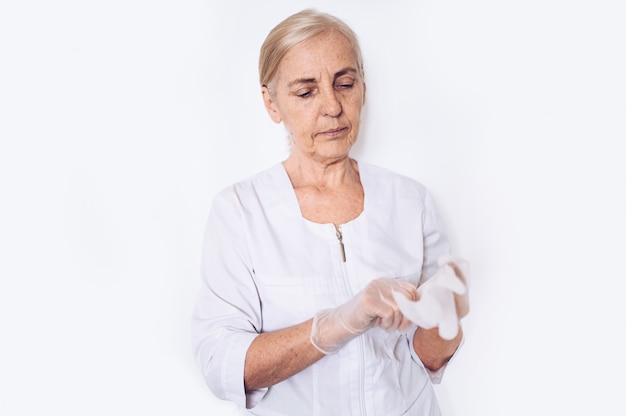 Elderly senior mature woman doctor or nurse in a white medical coat puts on gloves wearing personal protective equipment isolated. Healthcare and medicine concept. Covid-19 pandemic crisis