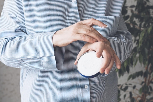 An elderly woman in a blue shirt applies cream to her hands