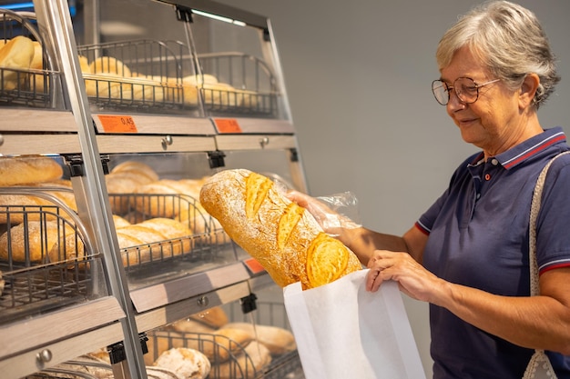 Elderly woman buying a rustic bakery bread at the supermarket by putting it in a paper bag