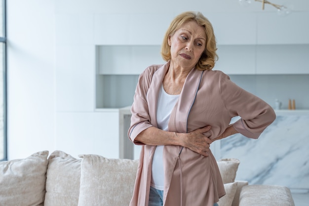 An elderly woman feels back pain while massaging aching muscles while standing in the living room