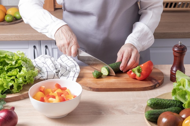 An elderly woman in a grey apron cuts a fresh green cucumber on a cutting board in a modern kitchen preparation of vegetable salad