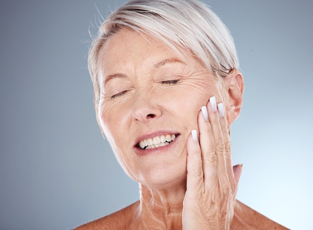 Elderly woman hand and smile for skincare cosmetics or moisturizer against a grey studio background Senior female touching face and smiling in satisfaction for healthy anti aging facial treatment