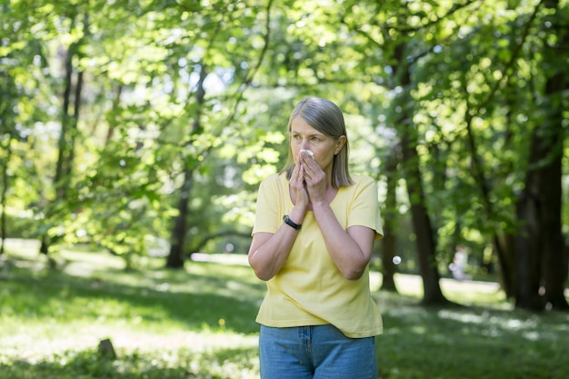 An elderly woman in the park with allergies has a runny nose and sneezes near trees