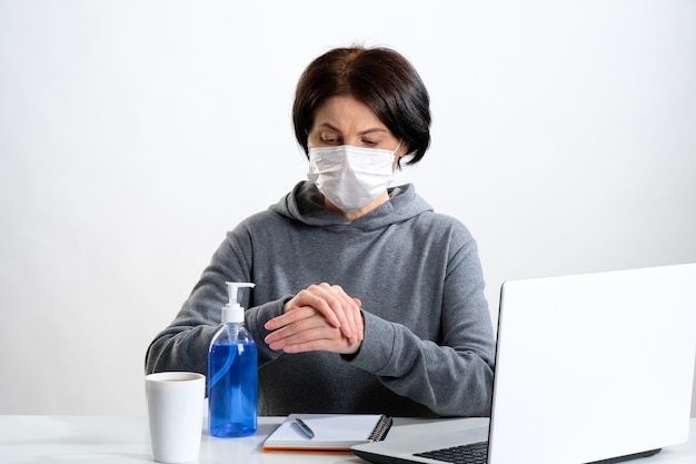 Elderly woman in a protective mask rubs her hands with antibacterial gel in the workplace