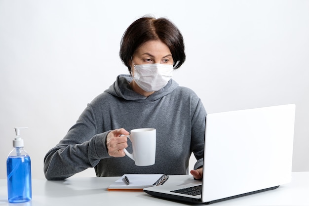 Elderly woman in a protective mask works at a computer and drinks coffee