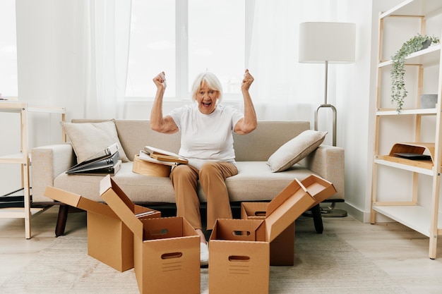 Photo elderly woman sits on a sofa at home with boxes collecting things with memories and moving and happiness smile