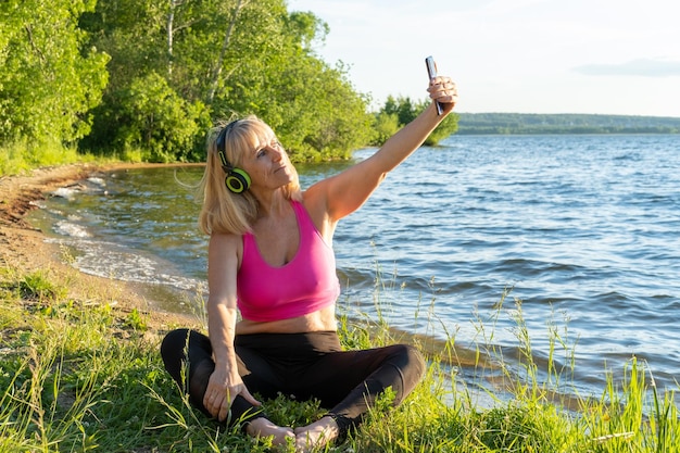 An elderly woman in sports clothes and headphones holds a phone in her hands watches online exercises for training waves a greeting with her hand listens to music takes selfies in a park