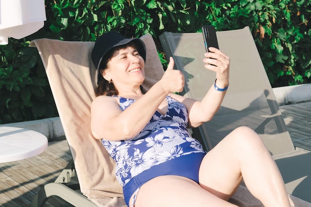 Elderly woman in swimsuit on sun lounger by the pool takes selfie on a smartphone and showing thumbs up