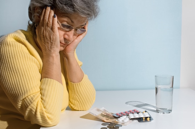 Elderly woman with glasses looking at money and pills on the table. Concept of depression, drug price, treatment cost