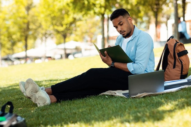 Elearning concept African american male student preparing for lecture using laptop and taking notes sitting in park