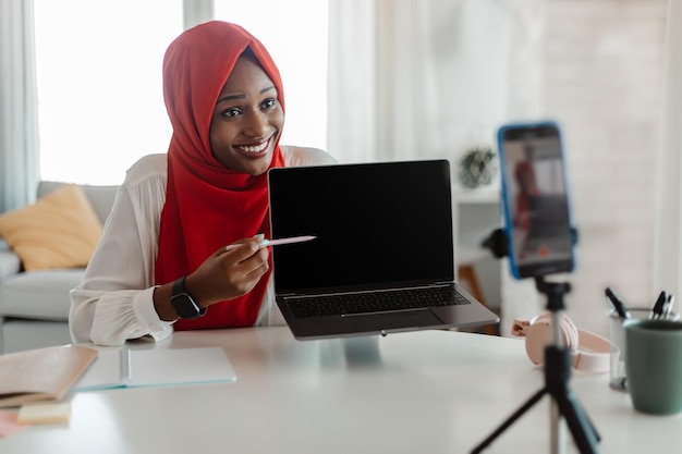 Elearning Positive black muslim woman having video call and showing blank laptop screen to cellphone camera