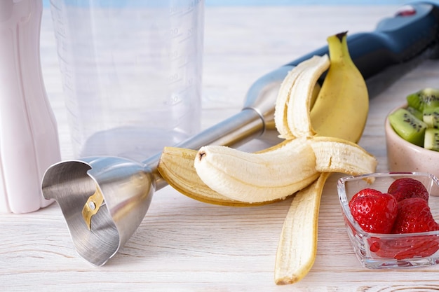 Electric hand blender with stainless steel body with accessories and fruits on wooden background, close-up.