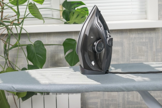 An electric iron on an ironing board on the background of a window with a green plant