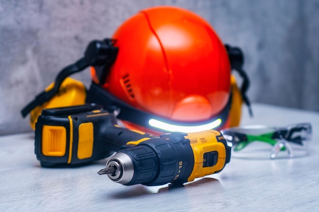 Electric screwdriver and a construction safety helmet for a head with an earpiece and a headlamp on the table Construction tool and form for protection