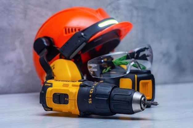 Electric screwdriver and a construction safety helmet for a head with an earpiece and a headlamp on the table Construction tool and form for protection